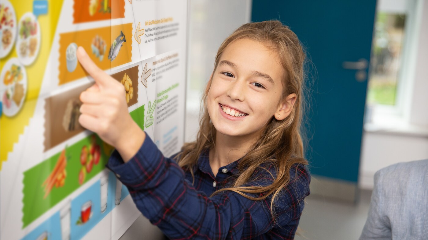 Ein Mädchen im Schulalter lacht und zeigt mit dem Zeigefinger auf ein großes Plakat mit der Ernährungspyramide..